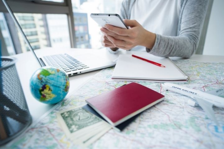 パソコンやパスポート、世界地図があるデスクでスマホを操作している手元を写した画像