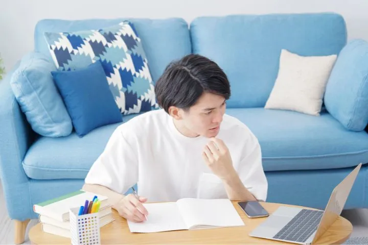 リビングで勉強している男性がノートパソコンを見つめて悩んでいる画像