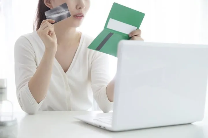 パソコンの前で通帳とカードを持っている人のイメージ