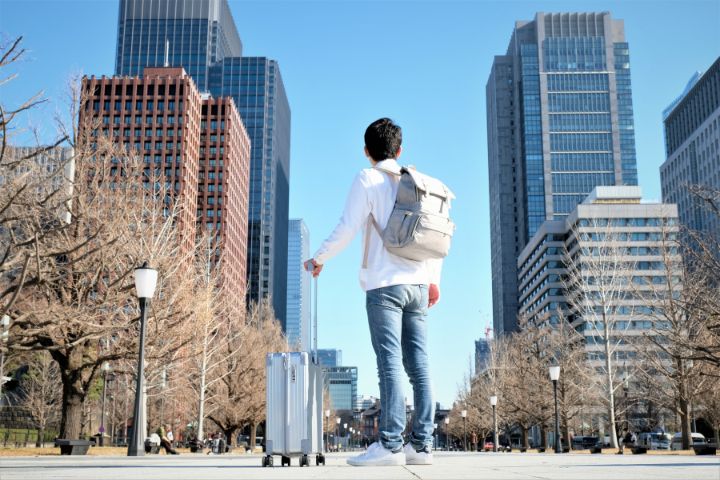 スーツケース片手にリュックを背負った男性がオフィス街を見ているイメージ