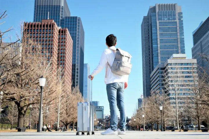 スーツケース片手にリュックを背負った男性がオフィス街を見ているイメージ