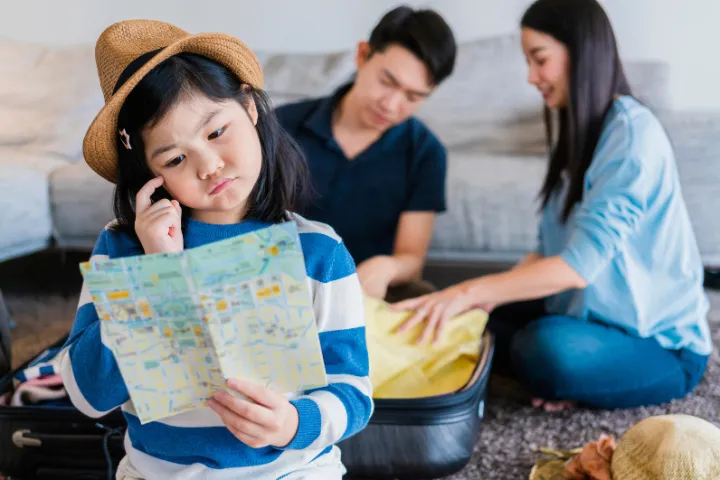 後ろで荷造りをする両親と地図を見て考えている女の子の画像