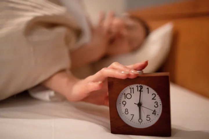 6時を差す目覚まし時計をあくびをしながら止める女性の画像