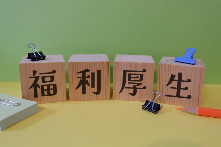 福利厚生と書かれた木のブロックとクリップと転がったオレンジの色鉛筆のイメージ