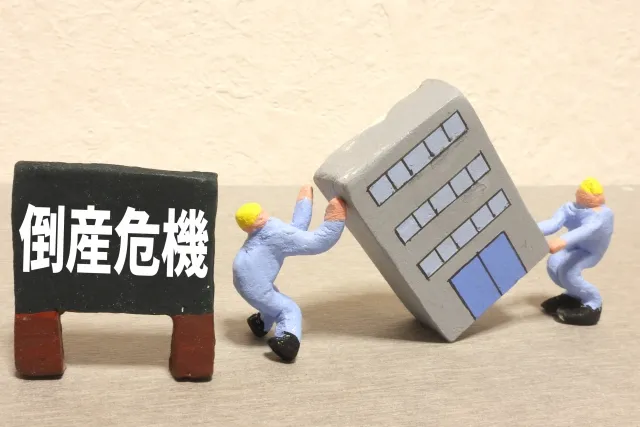 倒れるビルを押さえる人形と倒産危機と書かれた看板の画像