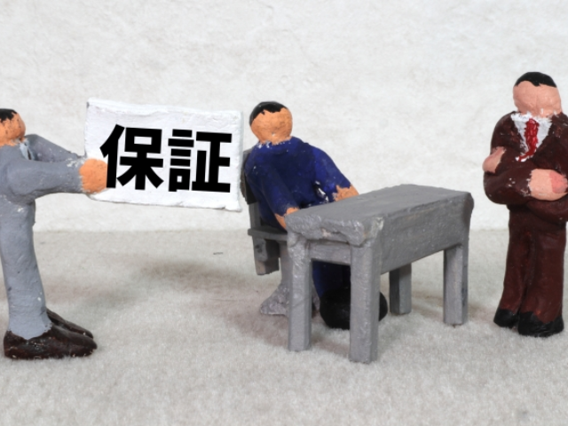 粘土でできたスーツ姿の人形が3体並んでおりそのうちの1体が保証と書かれた紙を持っている画像