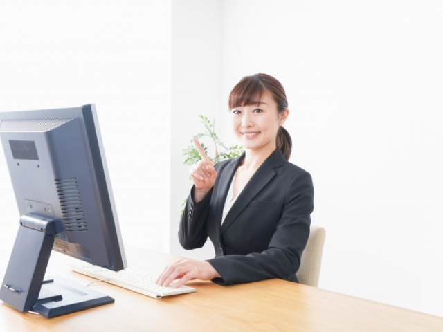 パソコンの前でスーツ姿の女性が笑顔で指差しをしている画像