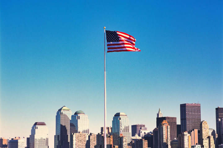 アメリカのビル群と青空の元で風にはためくアメリカ国旗の画像